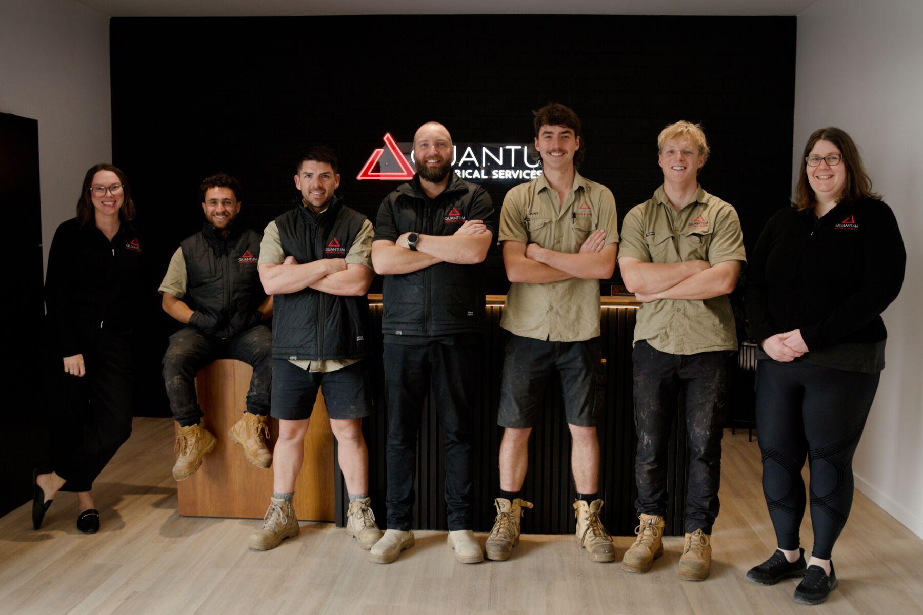 The Quantum Electrical Services team standing together in front of the illuminated Quantum sign at their office reception